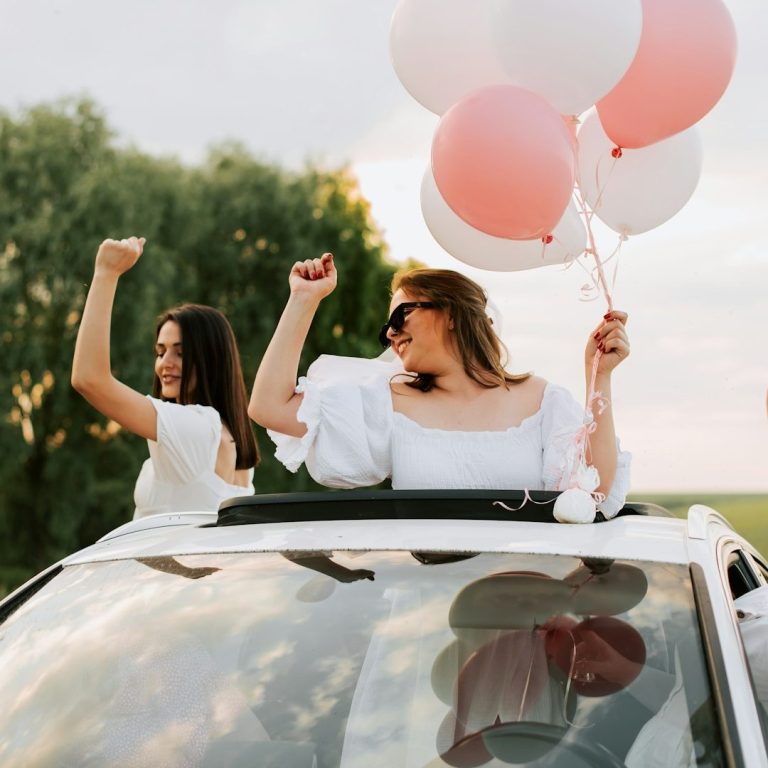 wedding balloons Two women celebrate in a car, holding pink and white balloons, with joyful expressions.